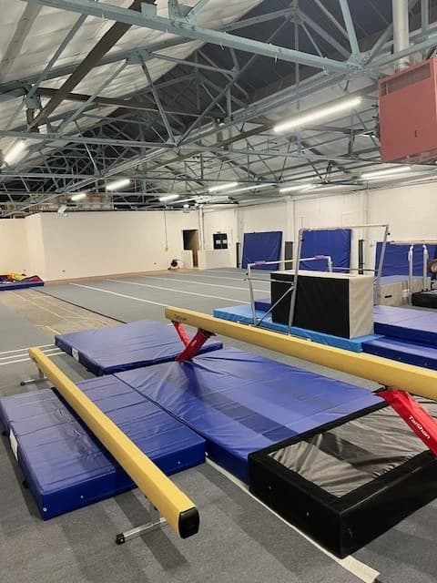 Two red aluminium FIG-approved competition balance beams with padded non-slip tops resting over blue landing mats in a gymnastics hall.