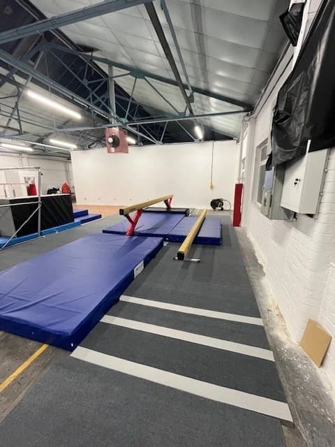 A red competition balance beam with tan top and red supports set on blue landing mats in a high-ceiling gym with metal trusses.