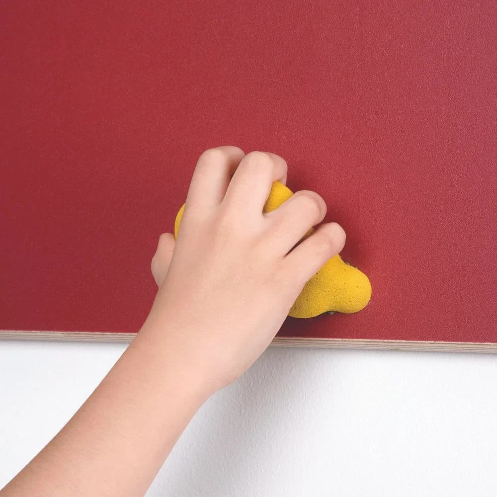 Hand gripping a yellow climbing hold mounted on a red traverse wall panel with a visible Finnish birch ply edge above a white wall.