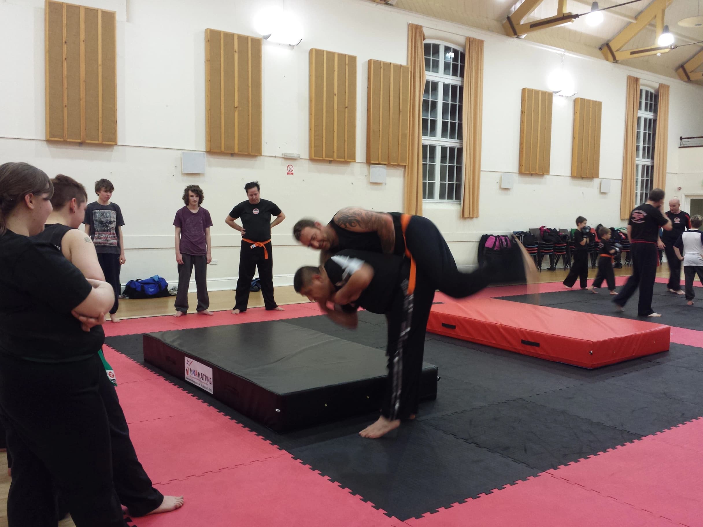 Two martial artists on a black and red mat in a gym, one throwing the other over his shoulder while classmates watch.