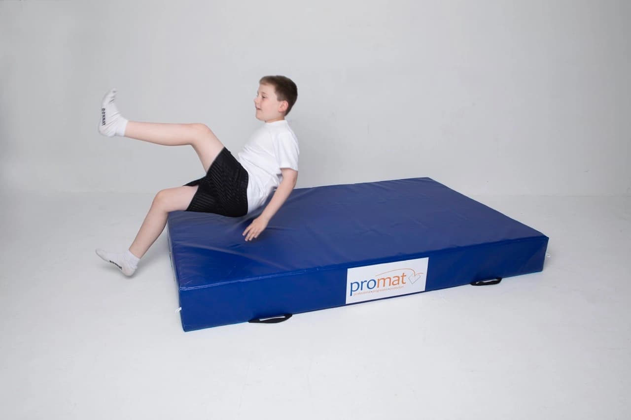 Boy in a white t-shirt, black shorts, and white socks sliding onto a blue mat labeled promat.