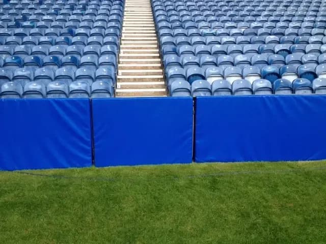 Blue padded barrier panels along the edge of a green pitch with rows of blue stadium seats and a central stairway.