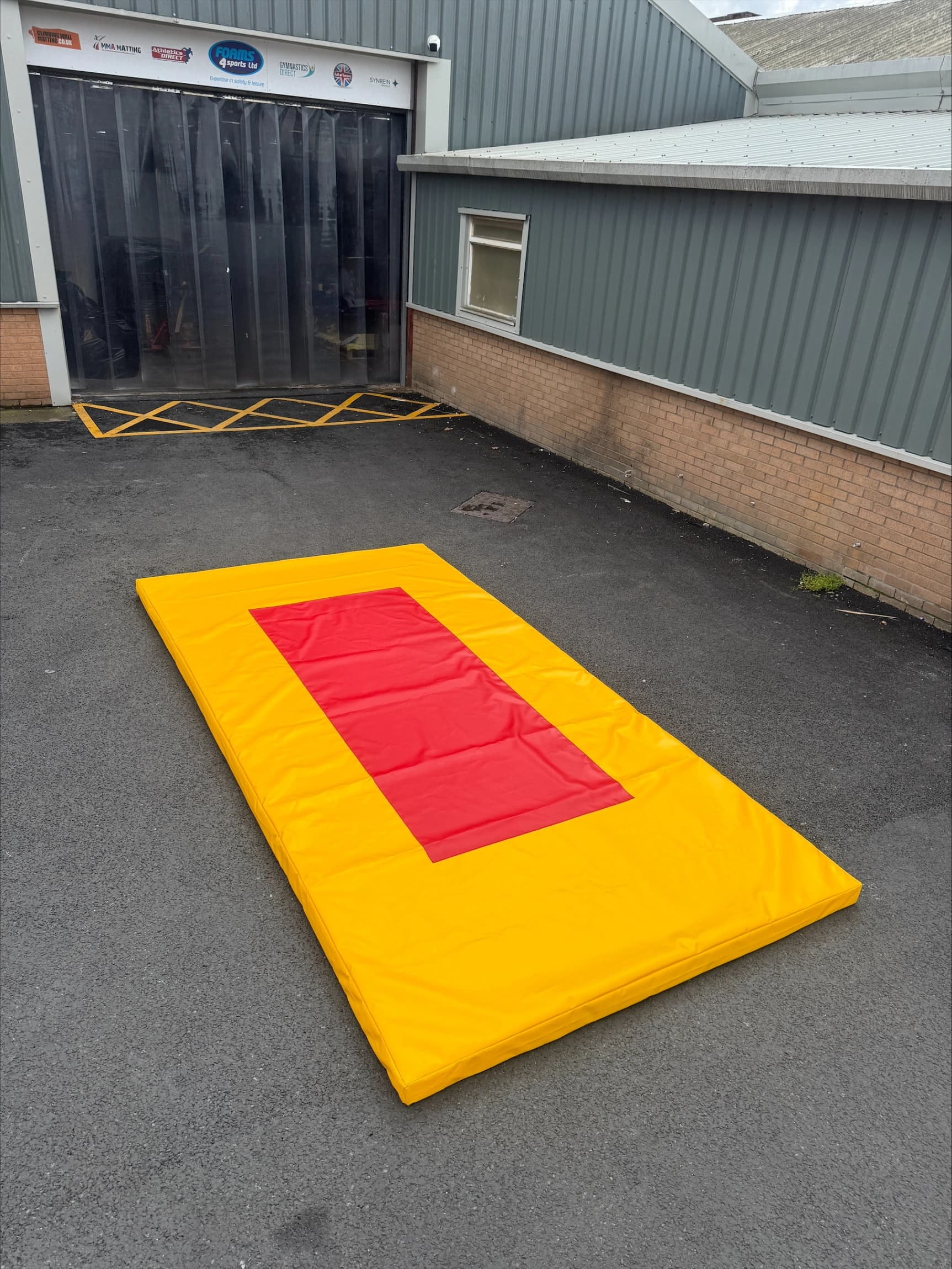 Bright yellow Stinger Mat with a central red rectangular target laid on asphalt outside a corrugated metal warehouse.