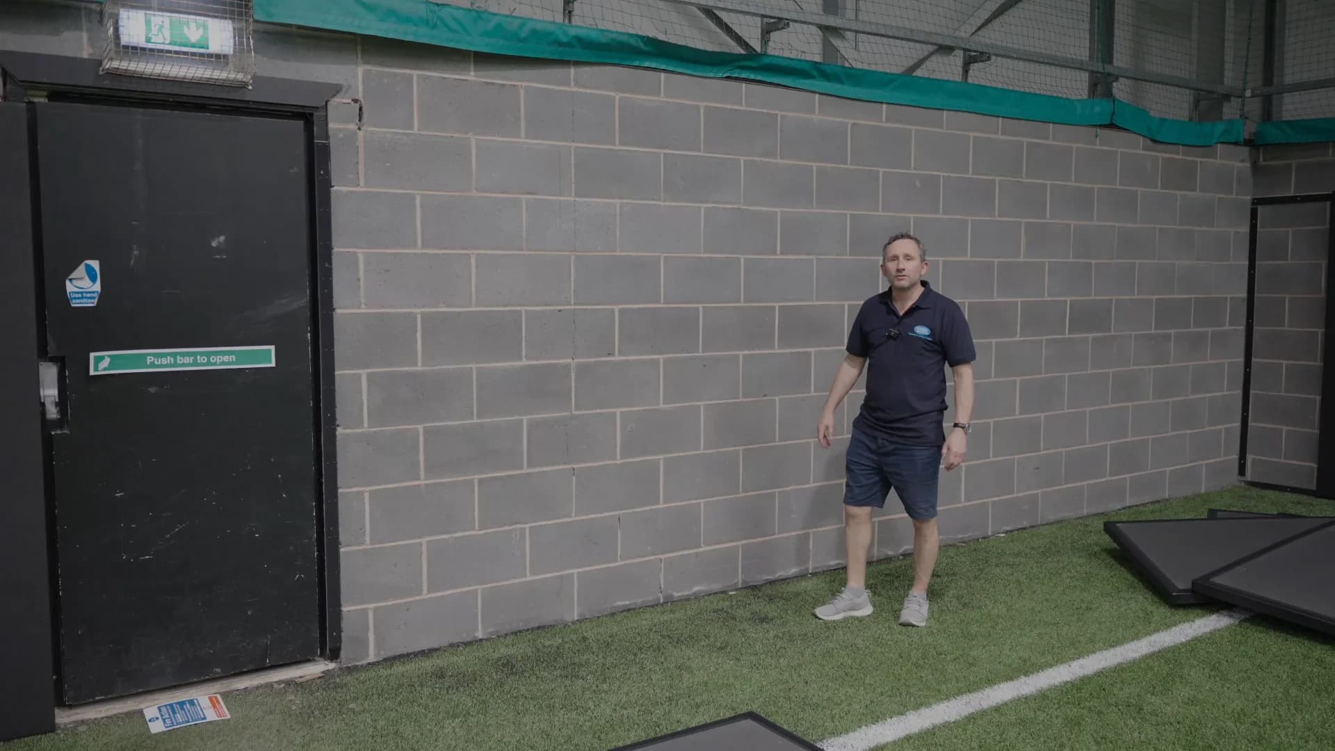 Man in a navy polo and shorts standing on indoor artificial turf beside a gray block wall and black door labeled "Push bar to open."